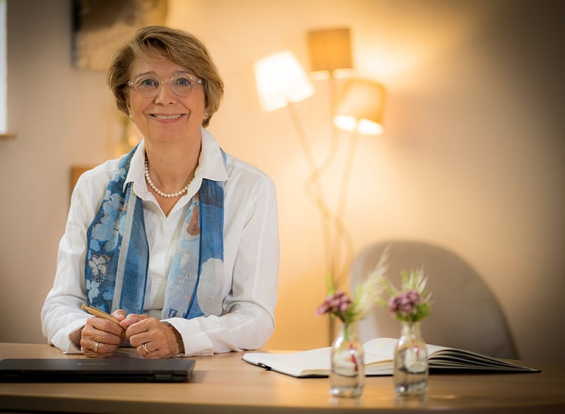 Daniela Sander-Lehmann hält einen Stift in der Hand und lächelt in die Kamera, im Hintergrund sind eine warme Lampe und zwei Pflanzen.